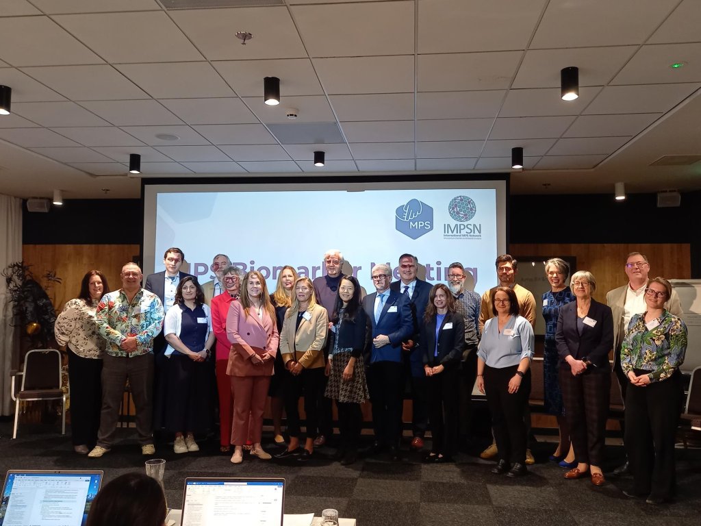 A diverse group of delegates stand in front of a projector screen with the MPS Society and IMPSN logos on it.